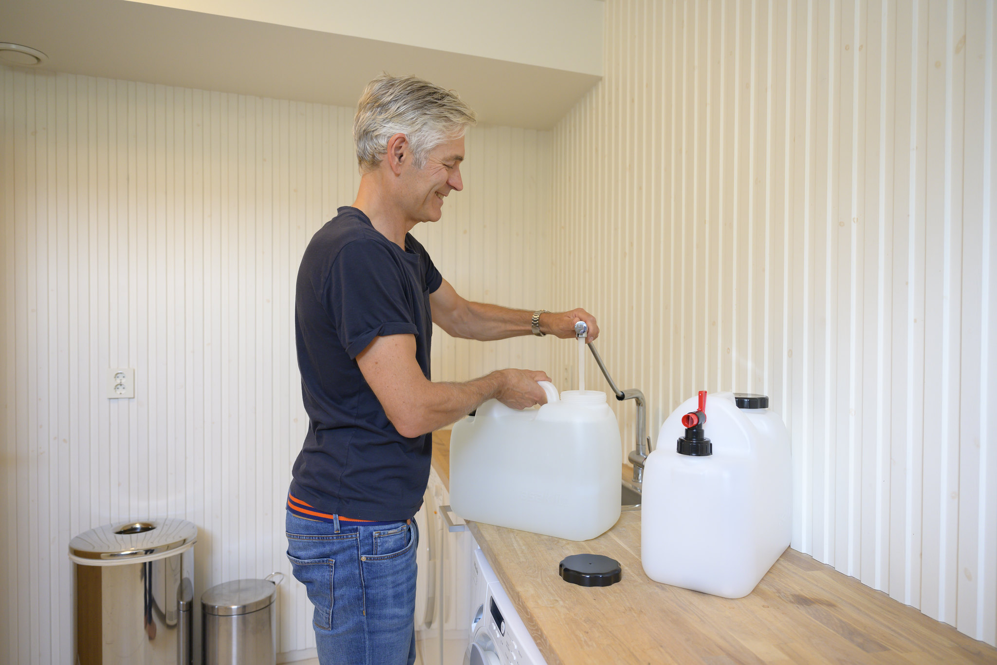 A man with gray hair and a dark T-shirt stands at a kitchen counter and fills two jugs with water.