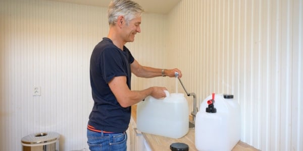 A man with gray hair and a dark T-shirt stands at a kitchen counter and fills two jugs with water.