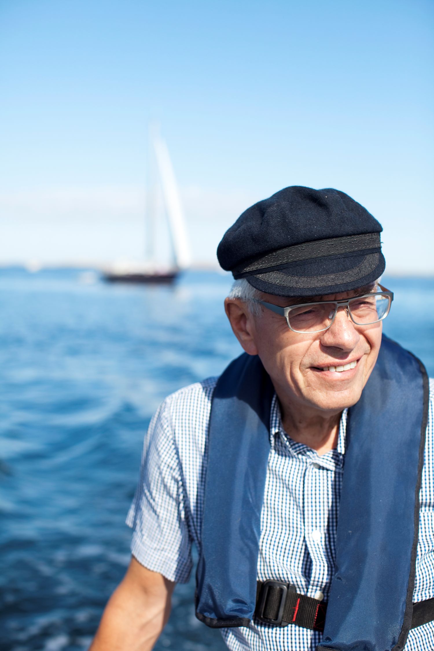 An elderly man with glasses, a hat and a short-sleeved shirt sits in a boat with an inflatable life jacket on. In the background, a sailboat can be glimpsed.