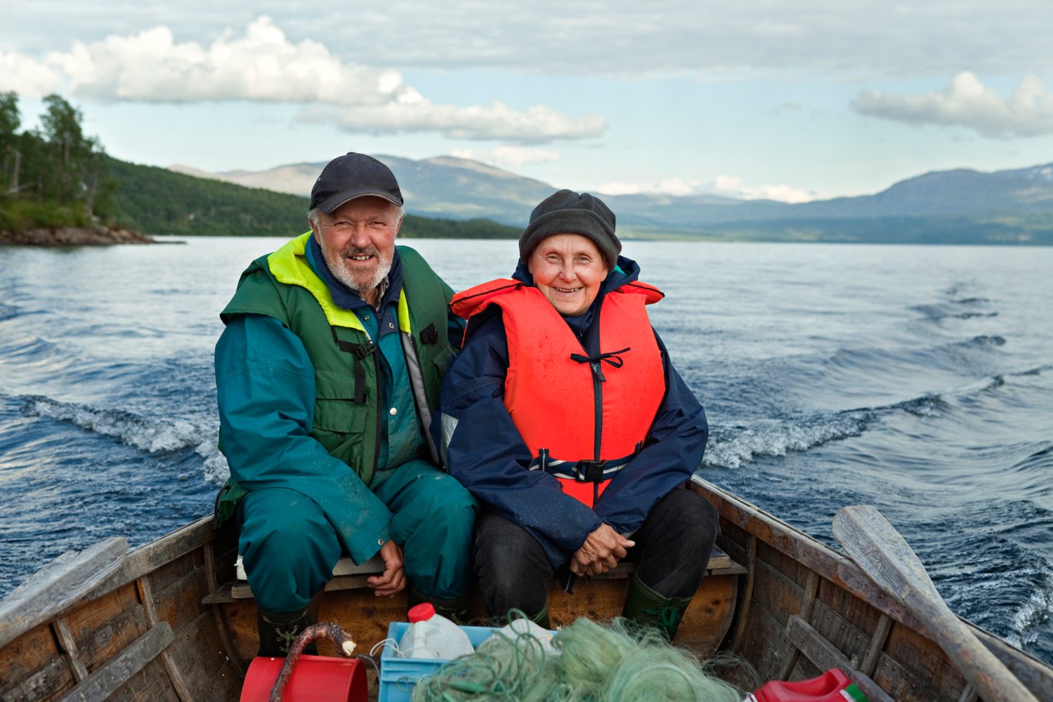 Eldre mann og kvinne sitter tett i en robåt av tre. Begge har flyteplagg på og smiler mot kamera.
