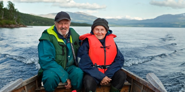 An elderly man and a woman sit close together in a wooden rowing boat. Both are wearing flotation suits and smiling at the camera.