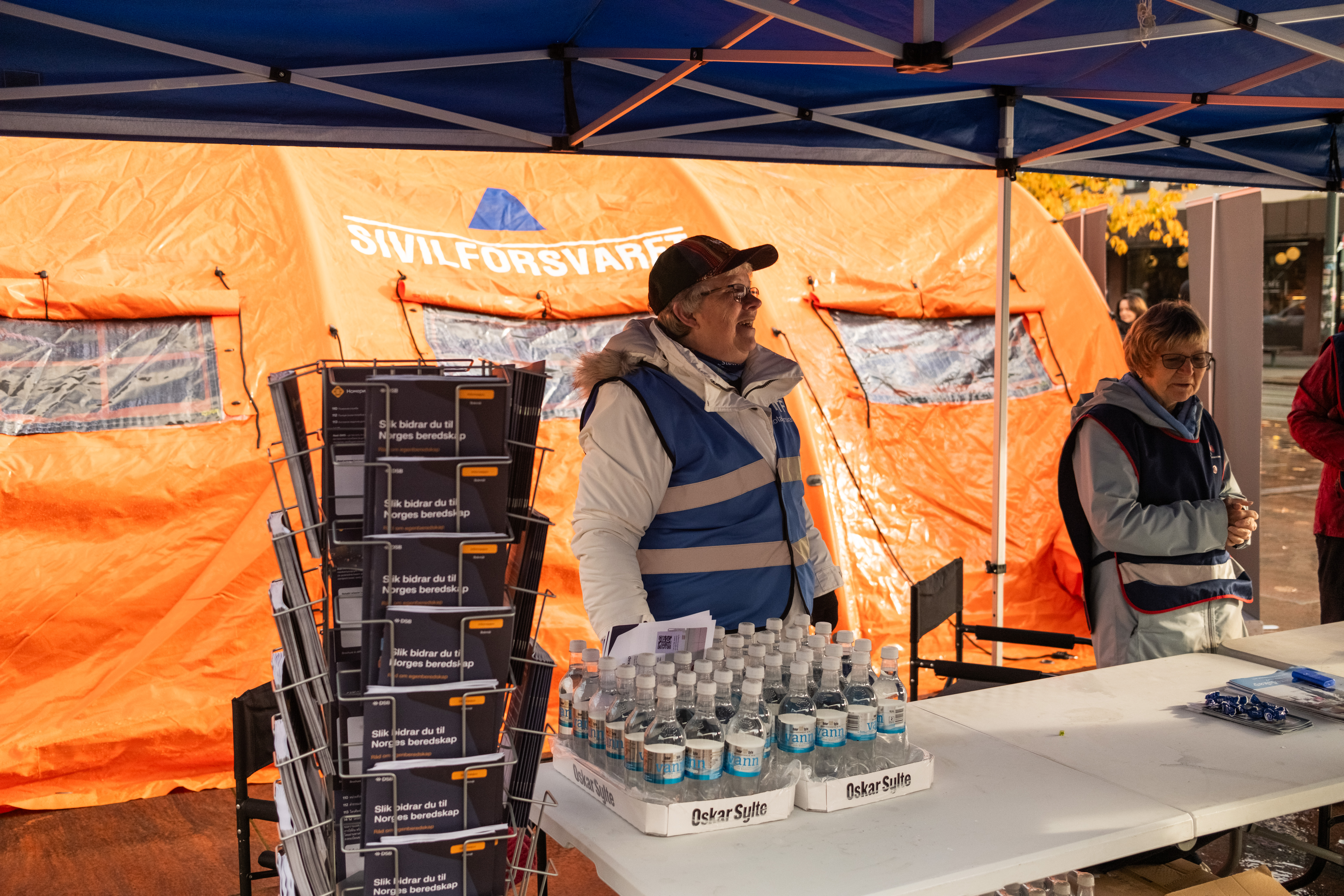 To personer med refleksvest som står bak et bord med vannflasker. I bakgrunnen skimtes et oransje sivilforsvarstelt.