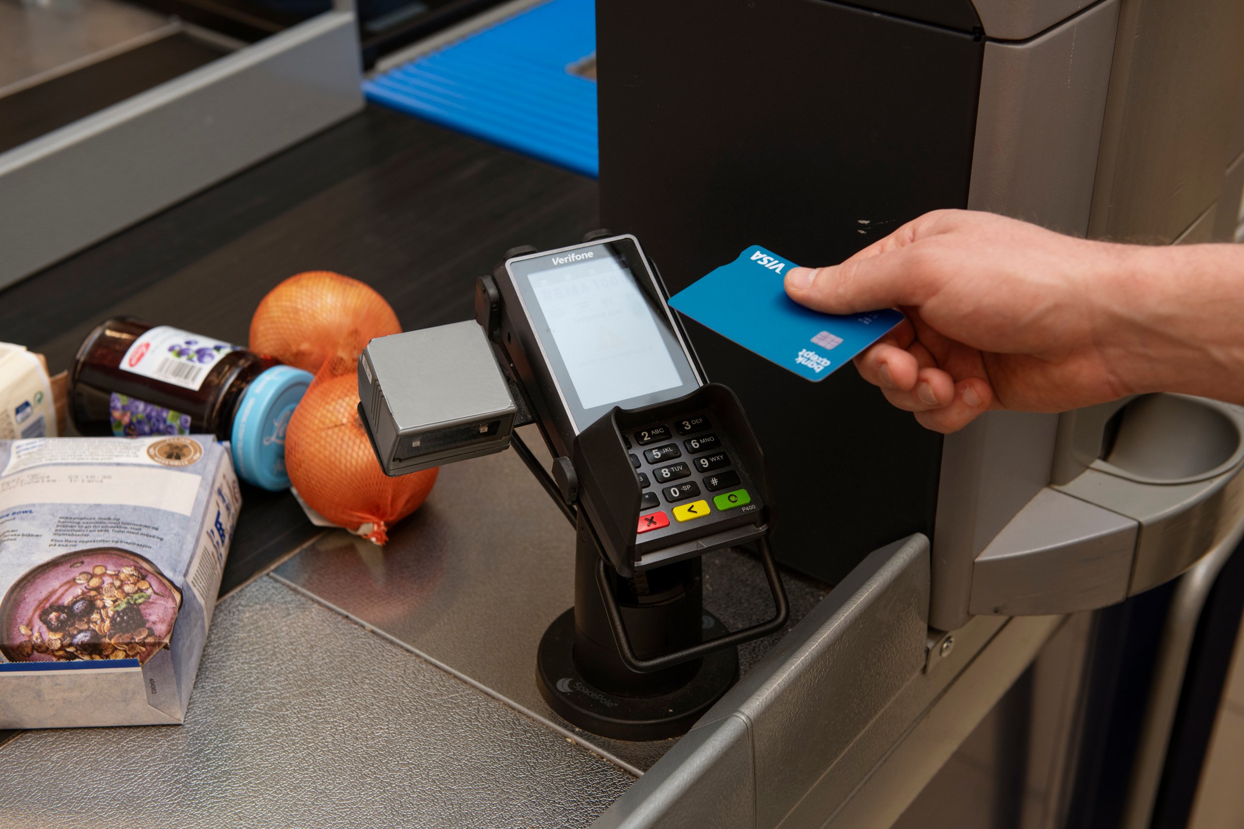 A hand holding a payment card against a payment machine. In the background, groceries can be seen on a shop counter.