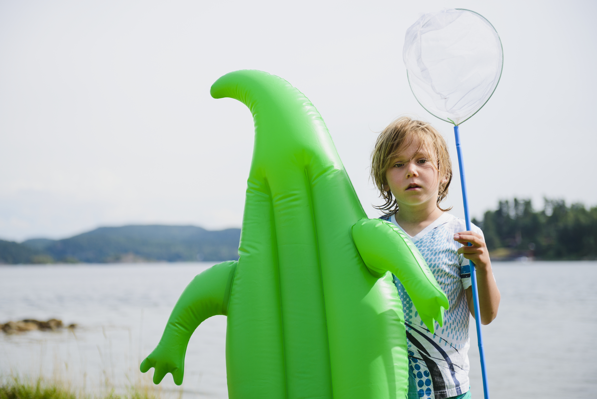 Boy with wet hair stands on the beach and holds up a large, inflatable green crocodile.