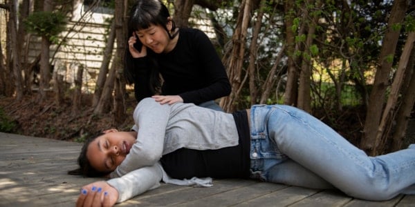 Ung kvinne som ligger med øynene lukket på et plankegulv utendørs. En ung kvinne sitter bak henne med hånden på skulderen hennes og snakker i mobiltelefonen.