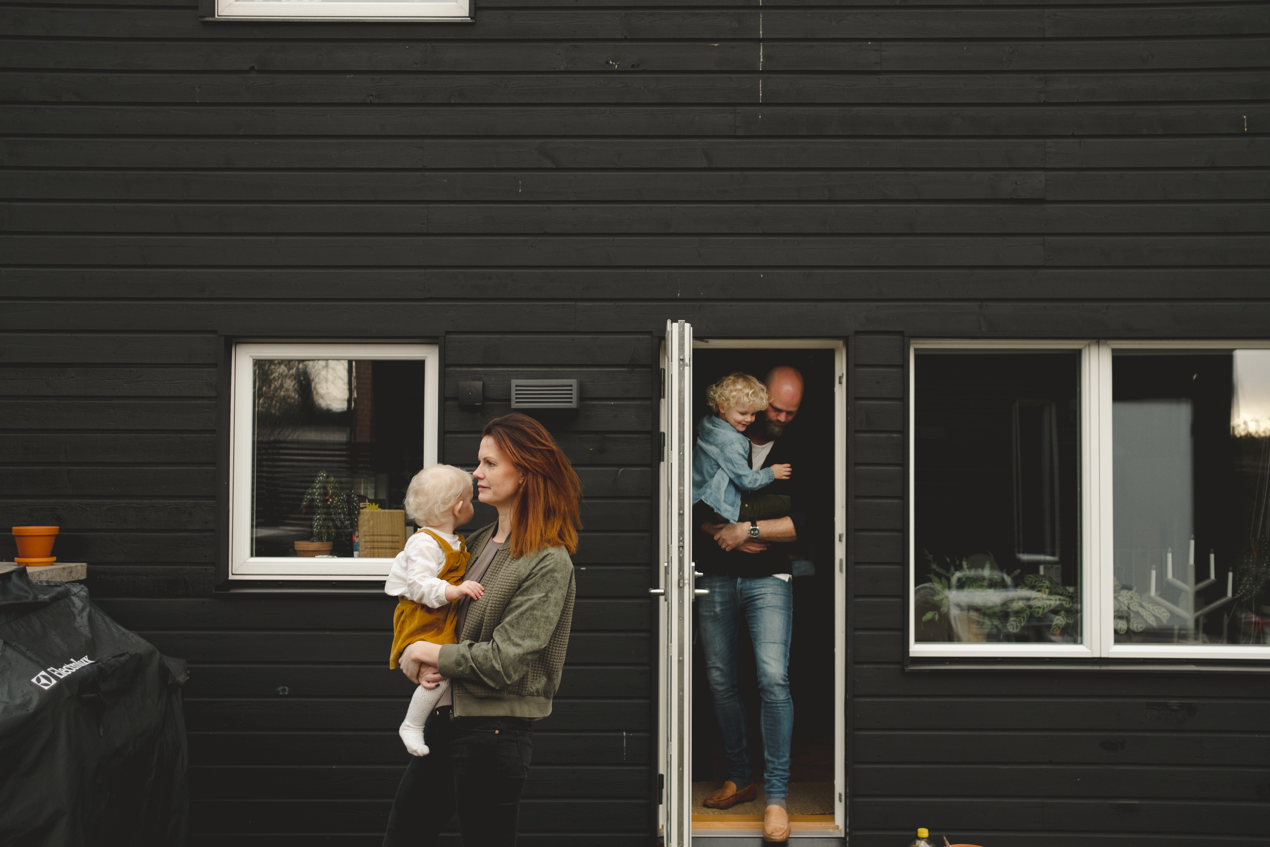 Mann og kvinne med hvert sitt barn på armen på vei ut fra en bolig. Huset er sort med hvite vinduskarmer.