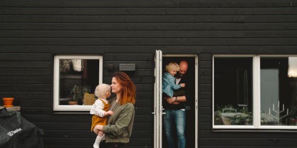 Mann og kvinne med hvert sitt barn på armen på vei ut fra en bolig. Huset er sort med hvite vinduskarmer.