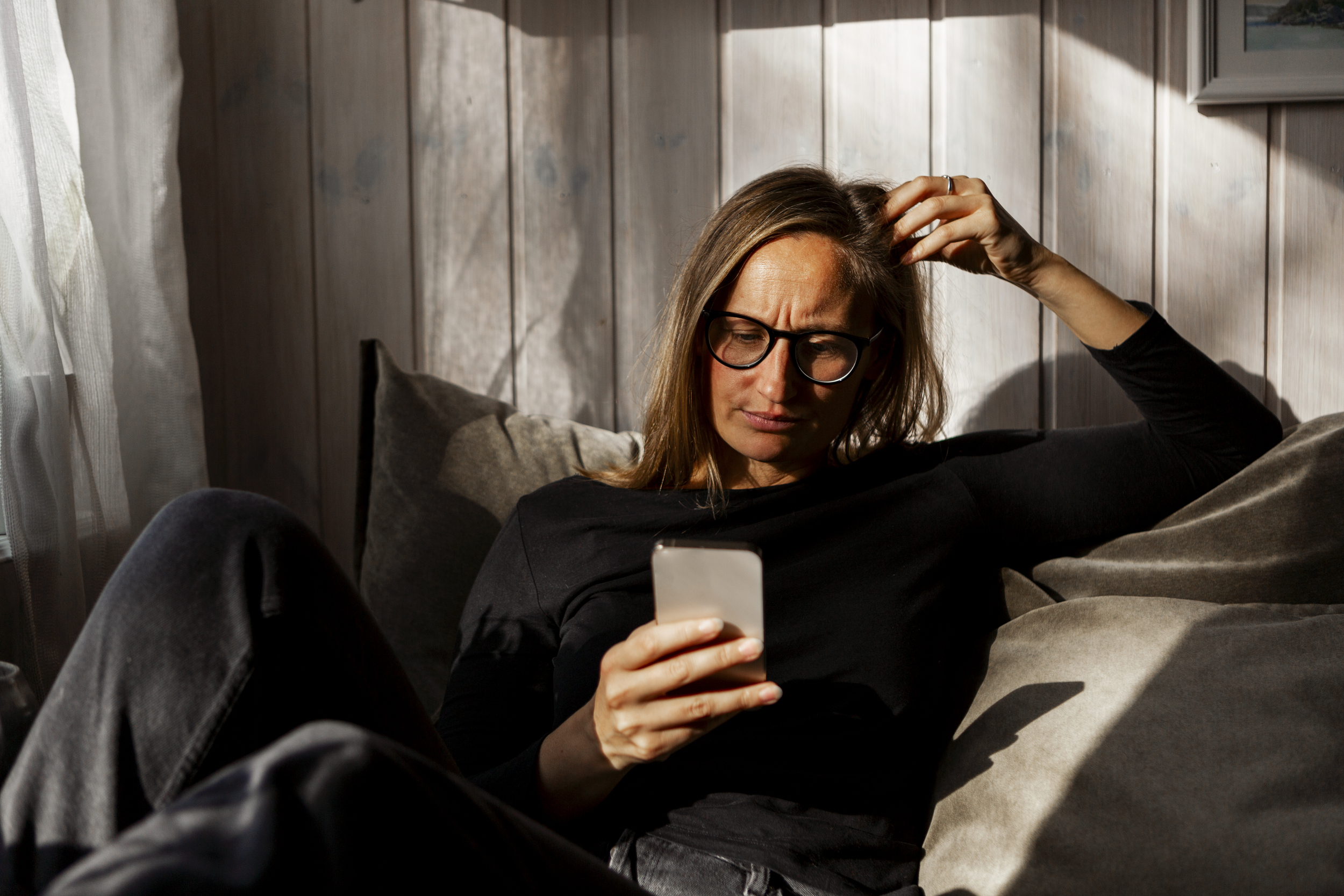 Kvinne med langt hår og briller sitter innendørs i halvmørke og ser på en mobiltelefon