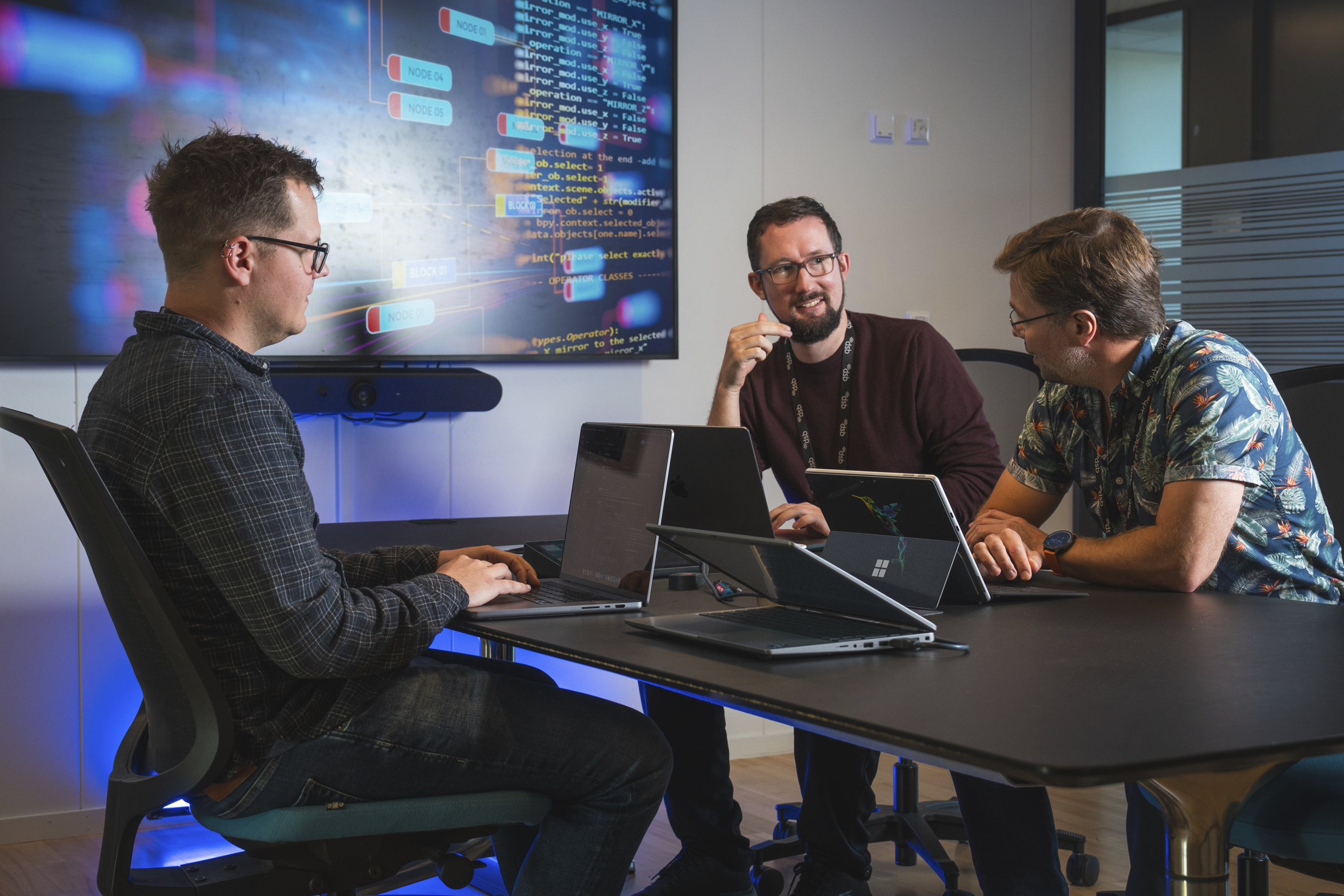 Tre menn som sitter og snakker sammen rundt et kontorbord. På bordet står flere bærbare pc'er.