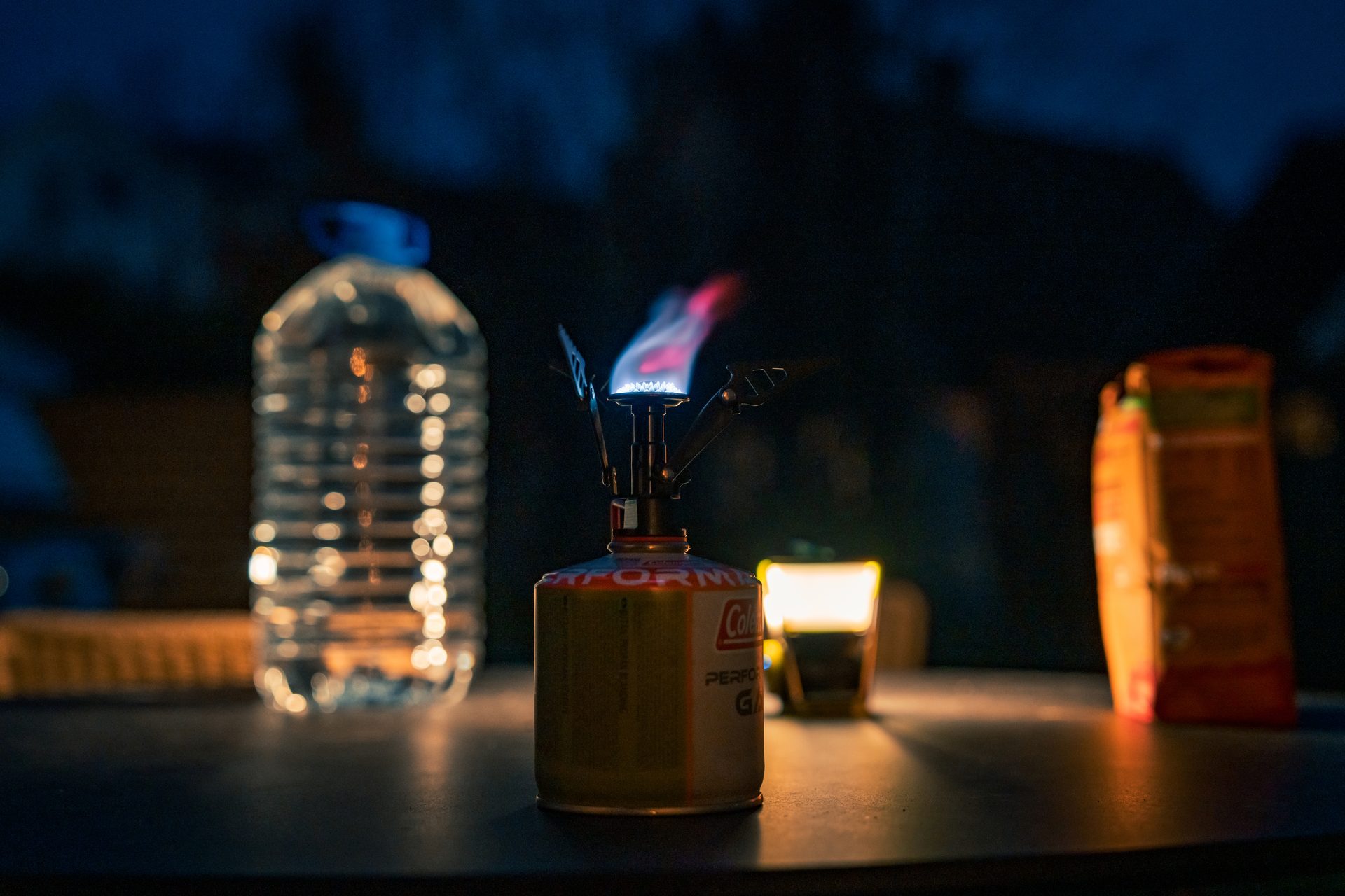 Lit camping stove, water bottle, and glowing lantern on a dark table at night.