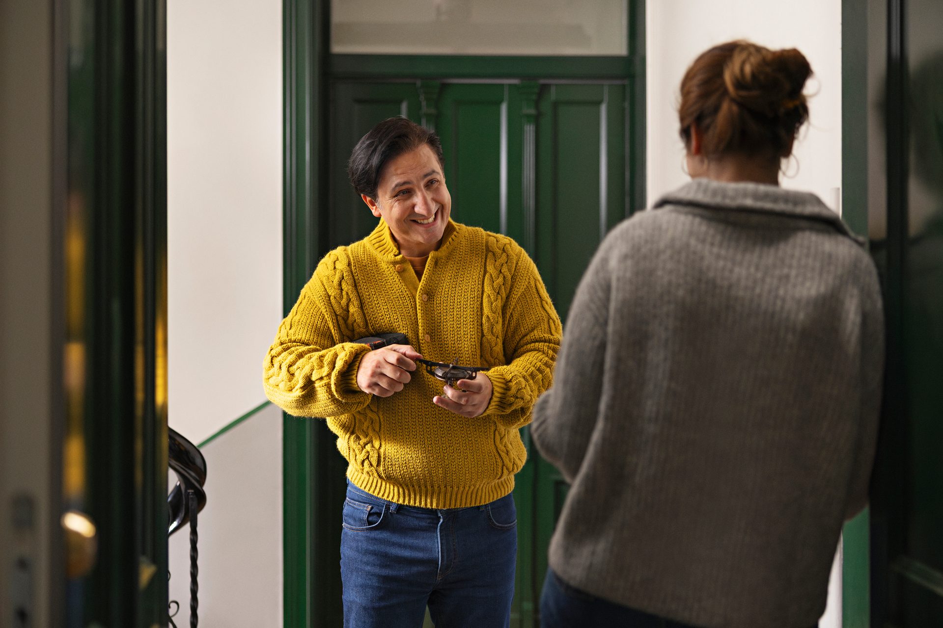 Smiling man in yellow sweater holding glasses talks to a woman in a doorway.
