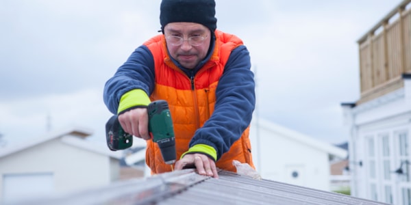 Mann med lue og arbeidsklær skrur fast takplate.
