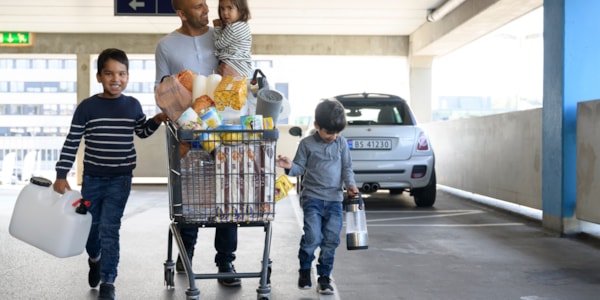 Man holding a little girl on his arm while pushing a shopping cart full of goods in front of him. On the side are two young boys carrying a watering can and a lantern.