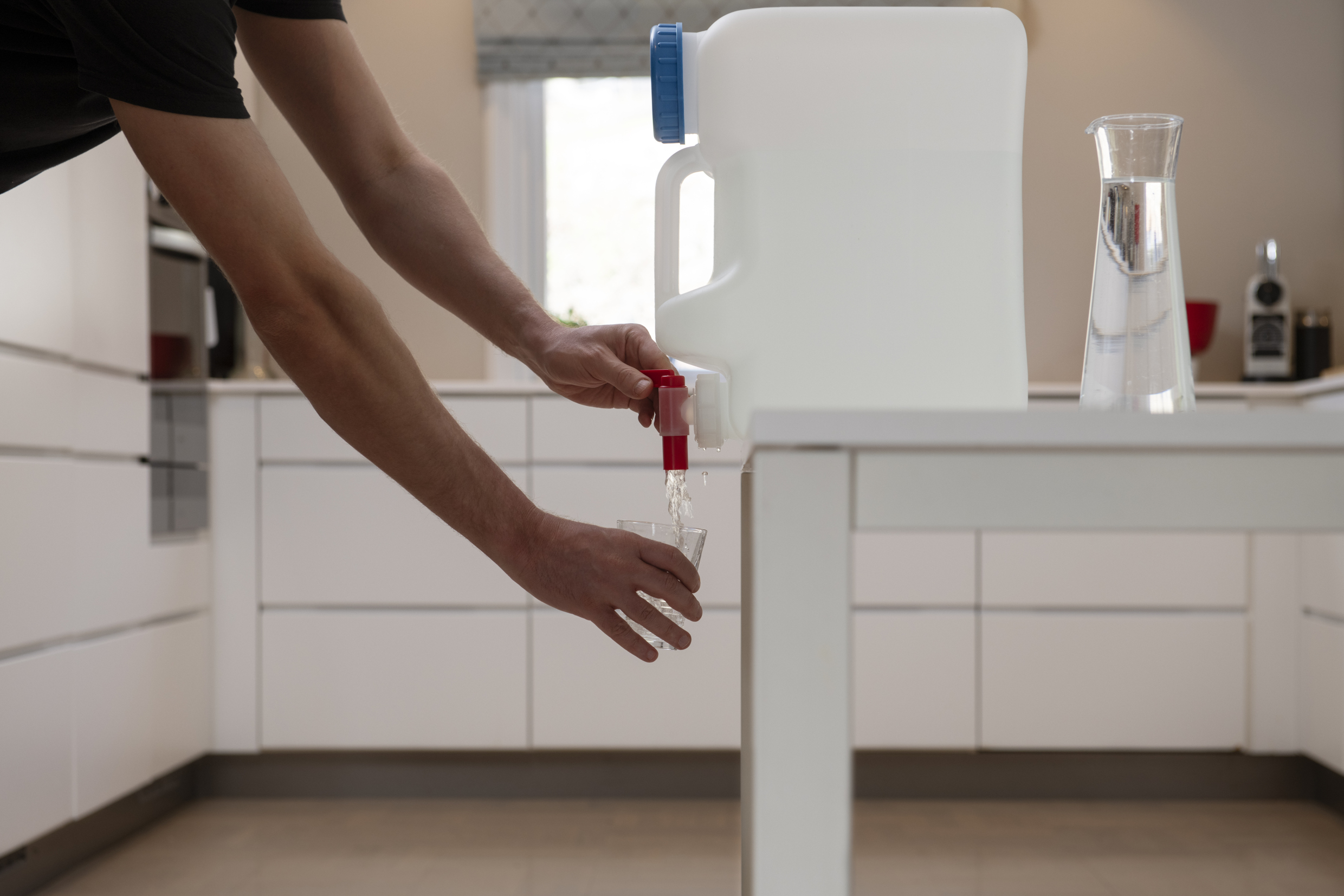 Cans of water standing on a table. A person whose arms we only see is draining water into a glass.