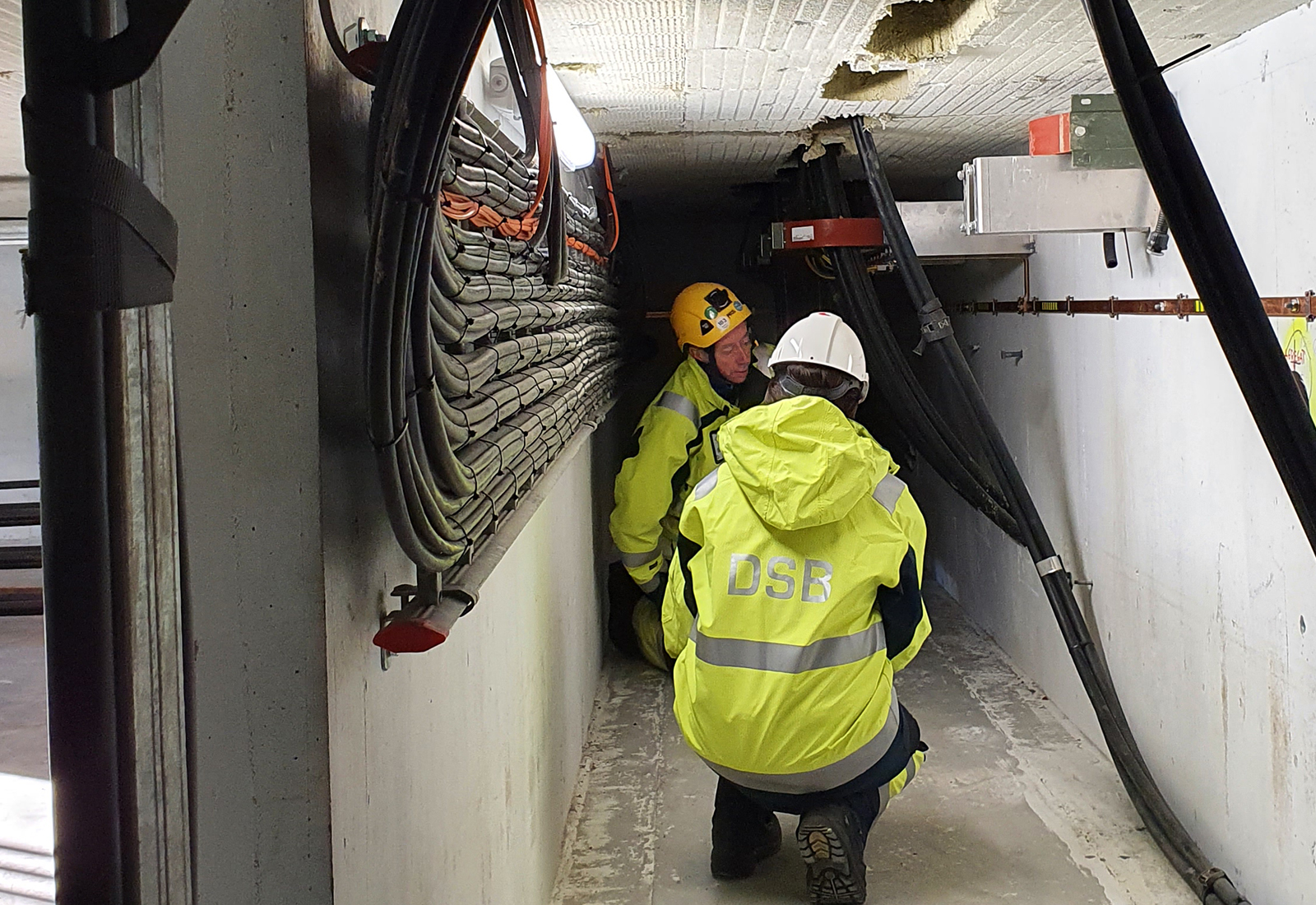To arbeidere i gule verneklær, med hjelm, inspiserer et teknisk rom med kabler