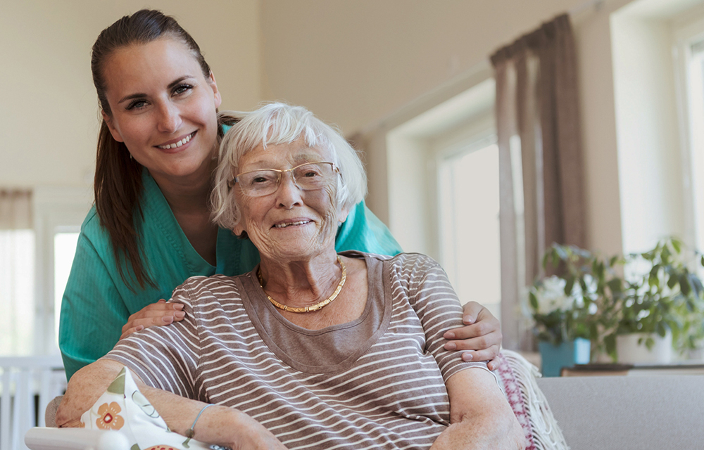 Kvinne i arbeidsuniform som står bak en eldre kvinne og holder henne på skuldrene. Begge smiler mot kamera.
