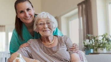 Kvinne i arbeidsuniform som står bak en eldre kvinne og holder henne på skuldrene. Begge smiler mot kamera.