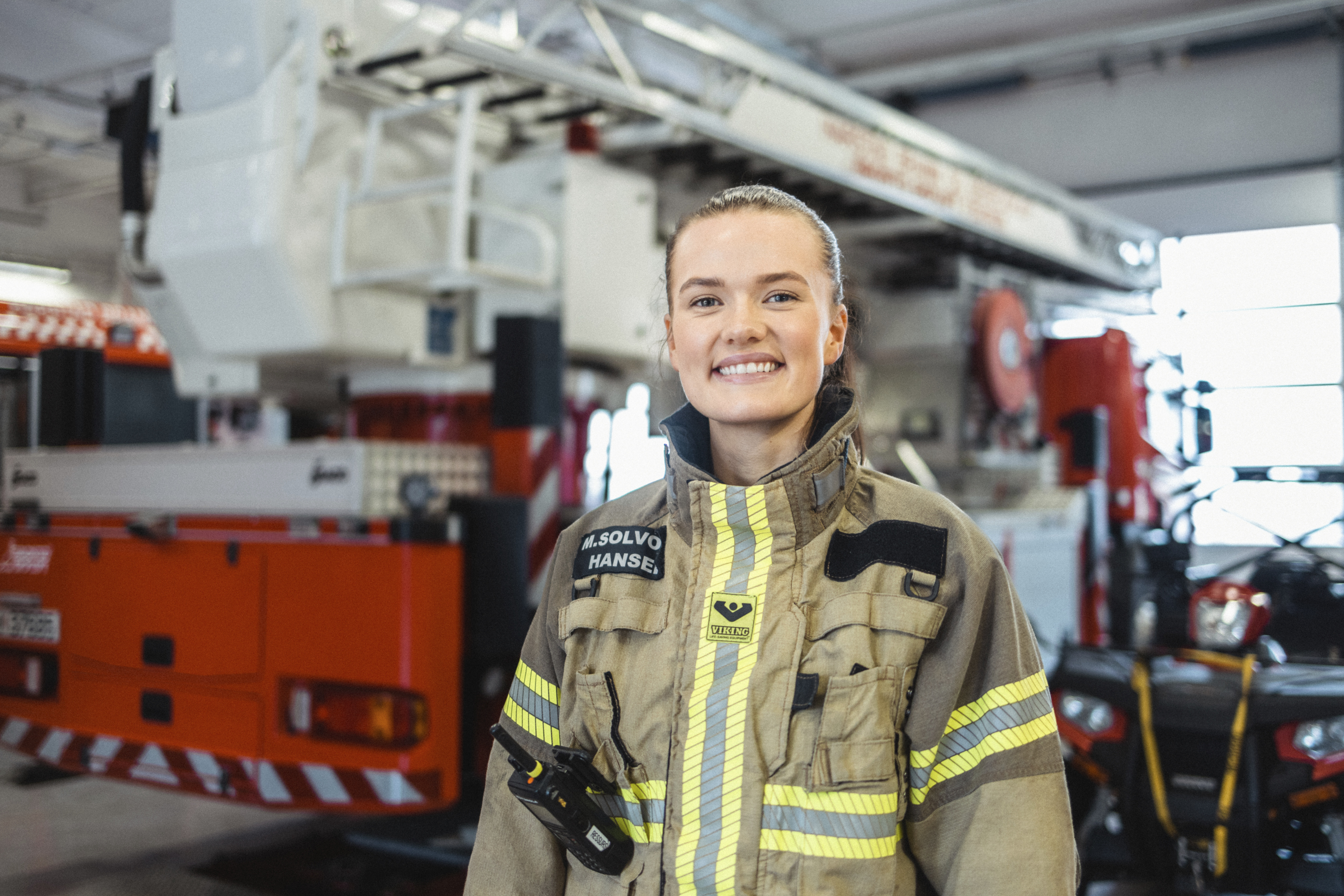 Ung kvinne i brannuniform smiler til kamera foran en brannbil.