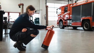 Person kneeling in a fire station preparing a fire extinguisher, with a fire truck in the background, representing emergency preparedness