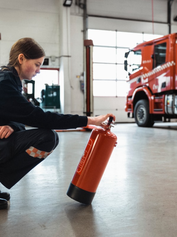 Person kneeling in a fire station preparing a fire extinguisher, with a fire truck in the background, representing emergency preparedness