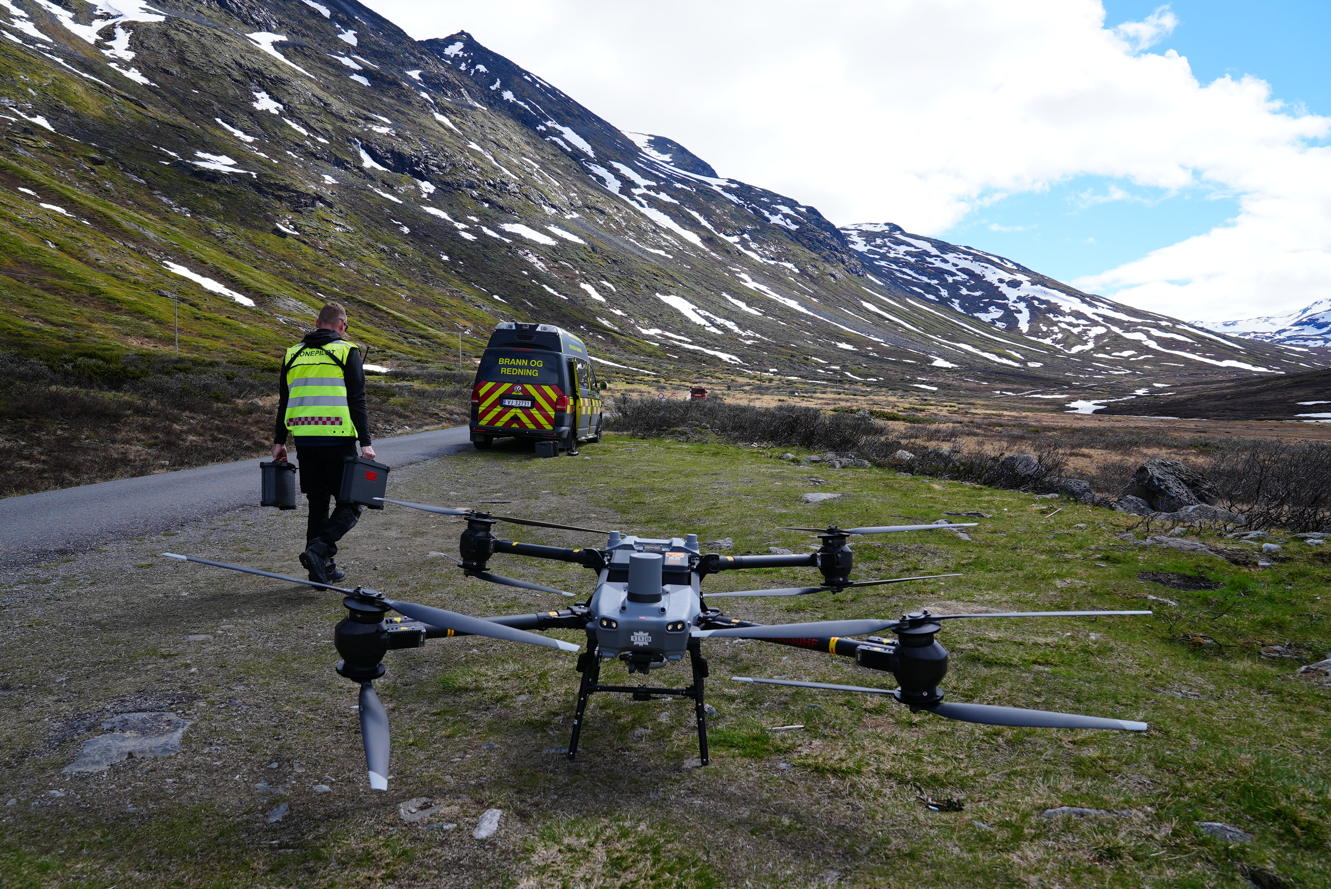 Drone og dronepilot i fjellandskap. I bakgrunnen står en varebil merket "Brann og redning"