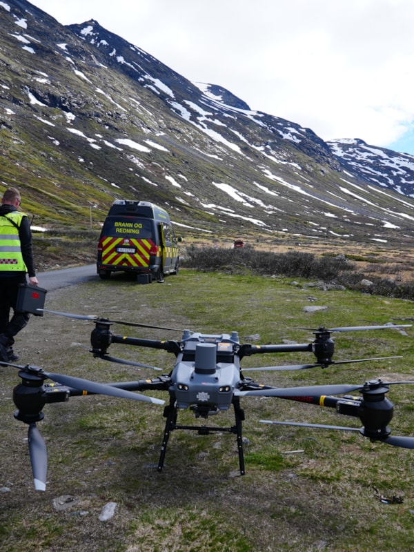 Drone og dronepilot i fjellandskap. I bakgrunnen står en varebil merket "Brann og redning"