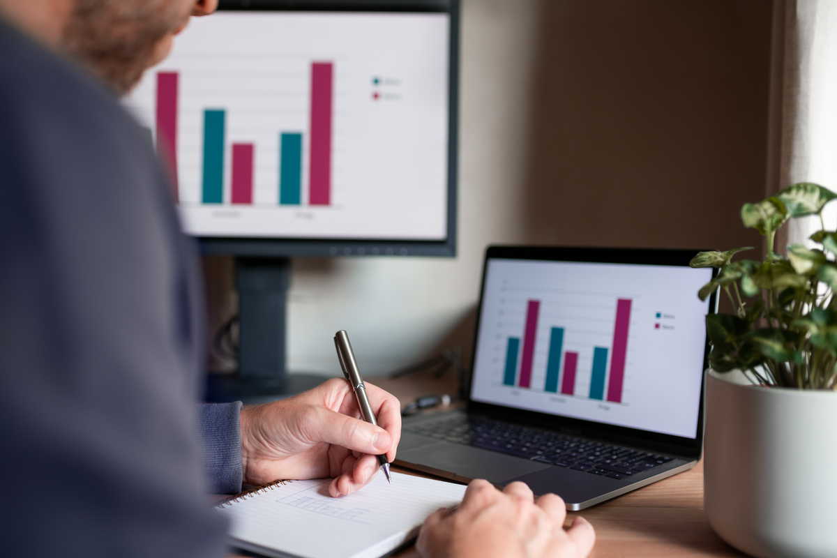 Halve kroppen til en mann som skriver på en notatblokk foran to skjermer med søylediagrammer