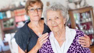 Kvinne med kort hår og briller holder rundt skuldrene på en eldre kvinne med grått, krøllete hår