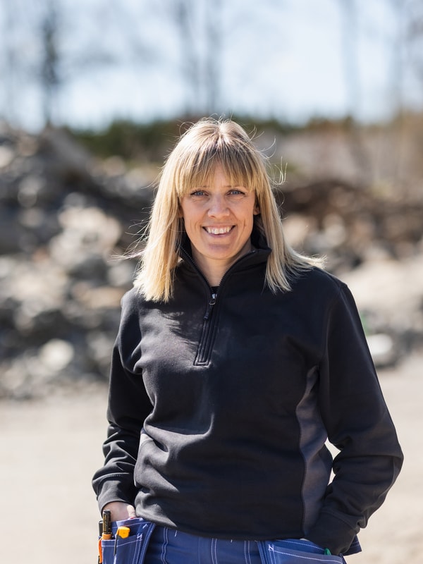 Person med blondt hår står utendørs foran en steinete bakgrunn iført sort genser og jeans, smiler mot kameraet under klar himmel