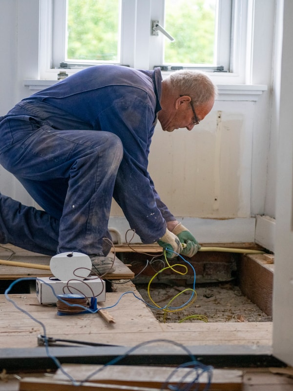Mann i kjeledress som sitter bøyd over et gulv og ordner med elektriske ledninger