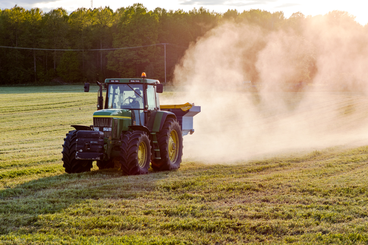 Traktor sprer gjødsel på åker i solnedgang, omringet av skog