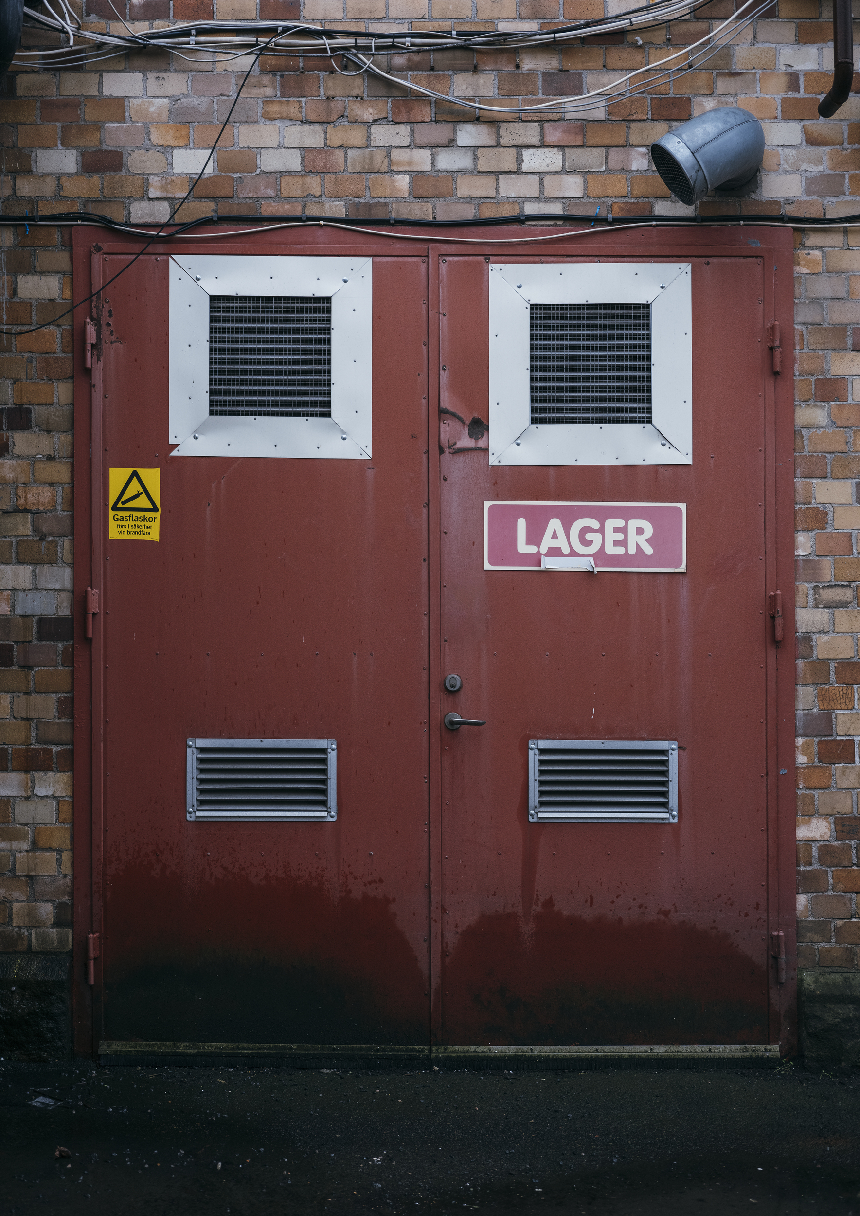 Rød dør i mursteinsvegg. På døren henger et skilt der det står "lager". Ved siden av henger et fareskilt for gass.