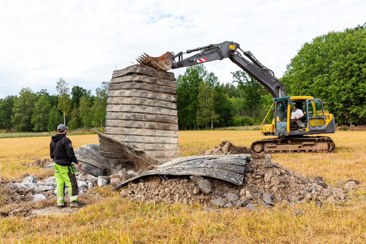 Gravemaskin river betongstruktur på åker, med en person som observerer, omgitt av trær