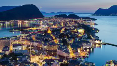 Flyfoto over Ålesund by om kvelden. Det lyser fra husene.