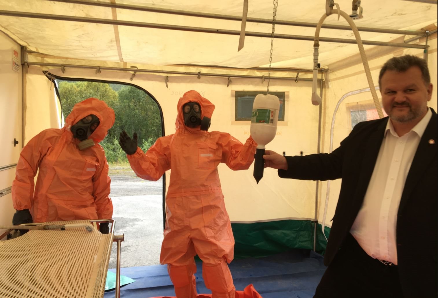 Personer ikledt oransje beskyttelsesdrakter inne i et telt sammen med en mann i dress.