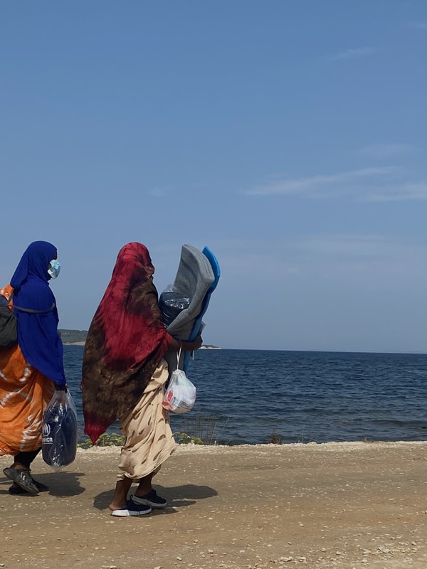 Kvinner i fargerike klær går langs en grusvei ved havet på en solrik dag, mens de bærer UNHCR-poser og tepper