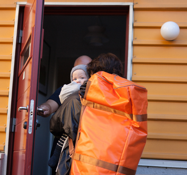 En person bærer et barn inn i et gult hus, mens en annen person holder døren åpen. Den bærende personen har på seg en stor oransje ryggsekk