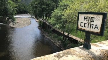 Gammelt skilt med teksten "Rio Ceira" på en betongkant. I bakgrunnen ses elven med trær og grønt gress langs elvebredden.