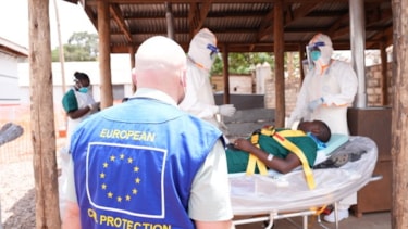 Norsk ekspert med EU-merket bekledning. I bakgrunnen ligger en mann på båre omgitt av personer med beskyttelsesdrakter.