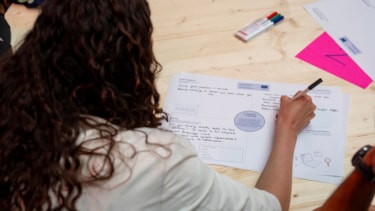 The back of the head of a woman with long hair writing on a piece of paper on a table