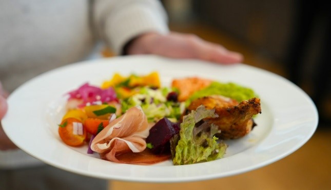 Hånd som holder frem en hvit tallerken med fargerik salat og spekeskinke