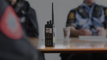 Nødnett-terminal som står på et bord. Rundt bordet sitter to politimenn og en brannmann.