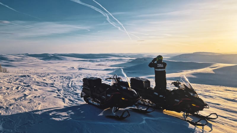 Tollinsektør med snøskuter speider med kikkert ut over snødekt landskap.