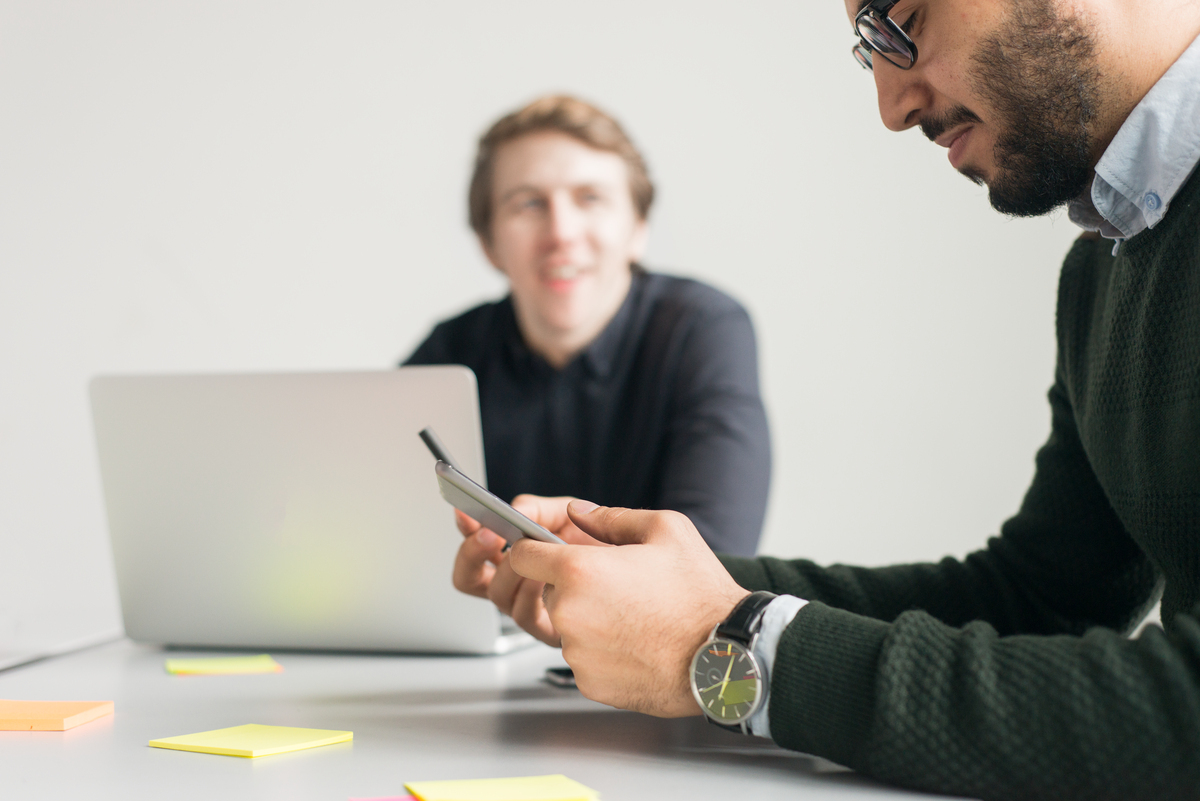 To mannlige kollegaer sitter ved et bord i et mterom. Den ene bak en pc og den andre med en mobiltelefon som han kikker ned på.