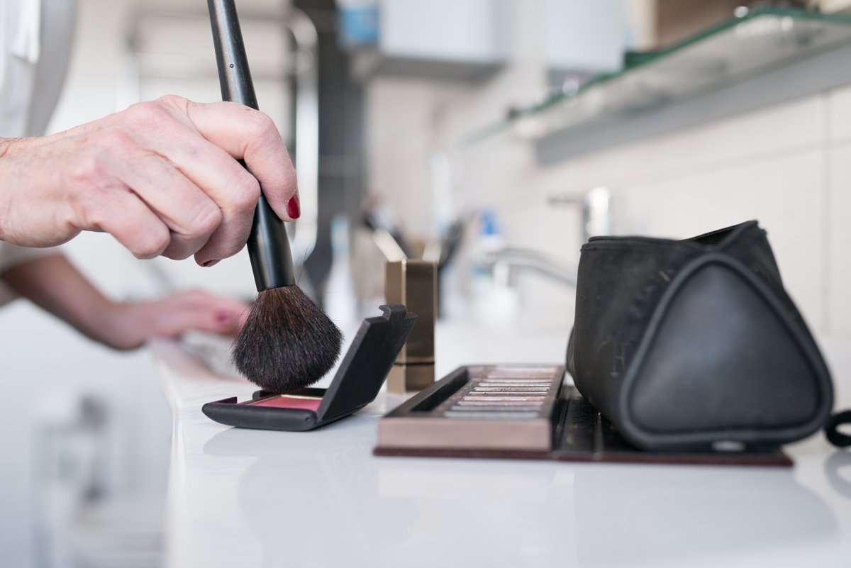 Person legger sminke med en børste ved siden av en sminkepalett og en svart sminkepose på en moderne baderomsvask