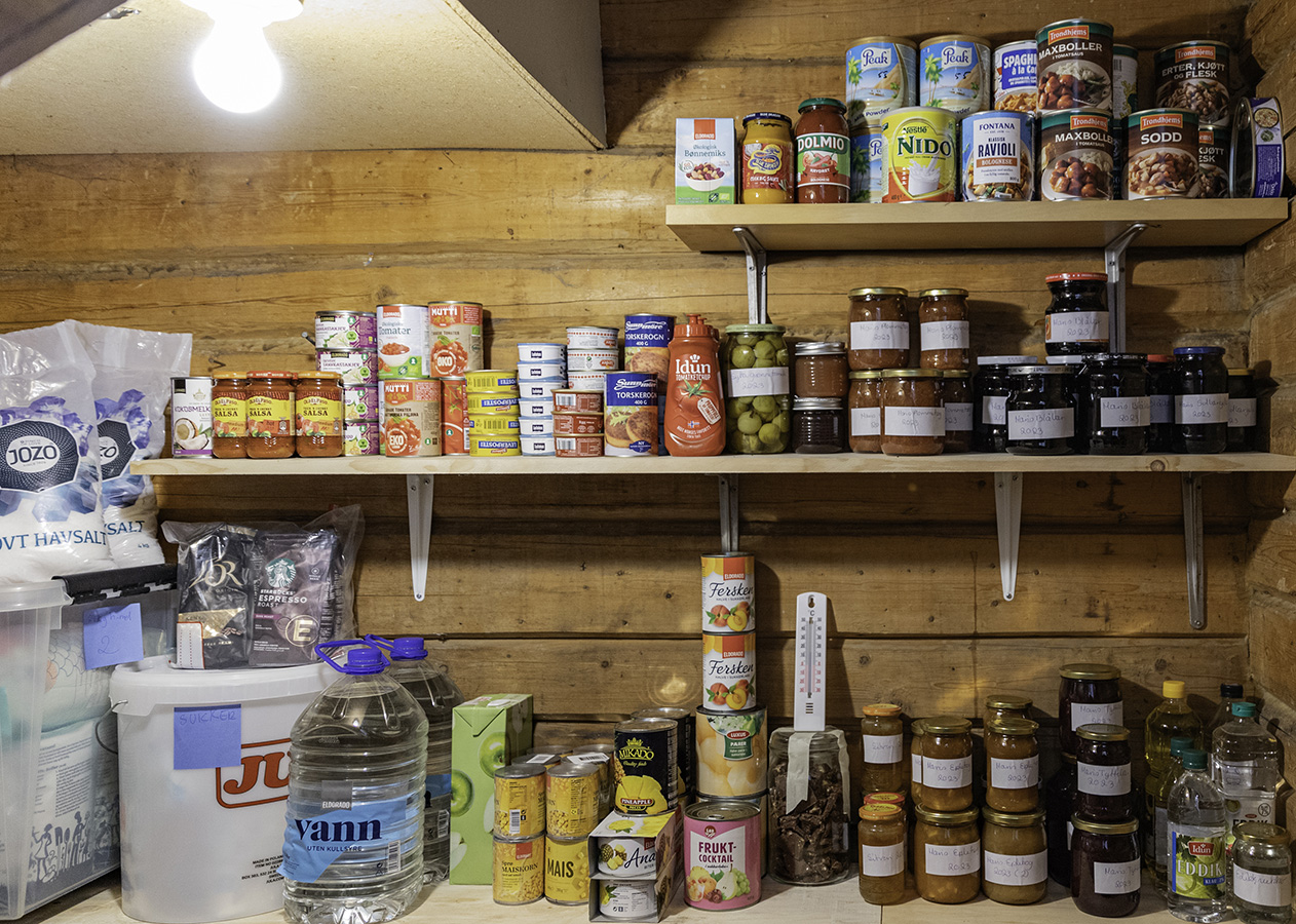 Hyller med vassdunker og diverse hermetikkboksar og syltetløyglass inne i ein bod. 