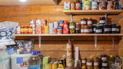 Hyller med vassdunker og diverse hermetikkboksar og syltetløyglass inne i ein bod.