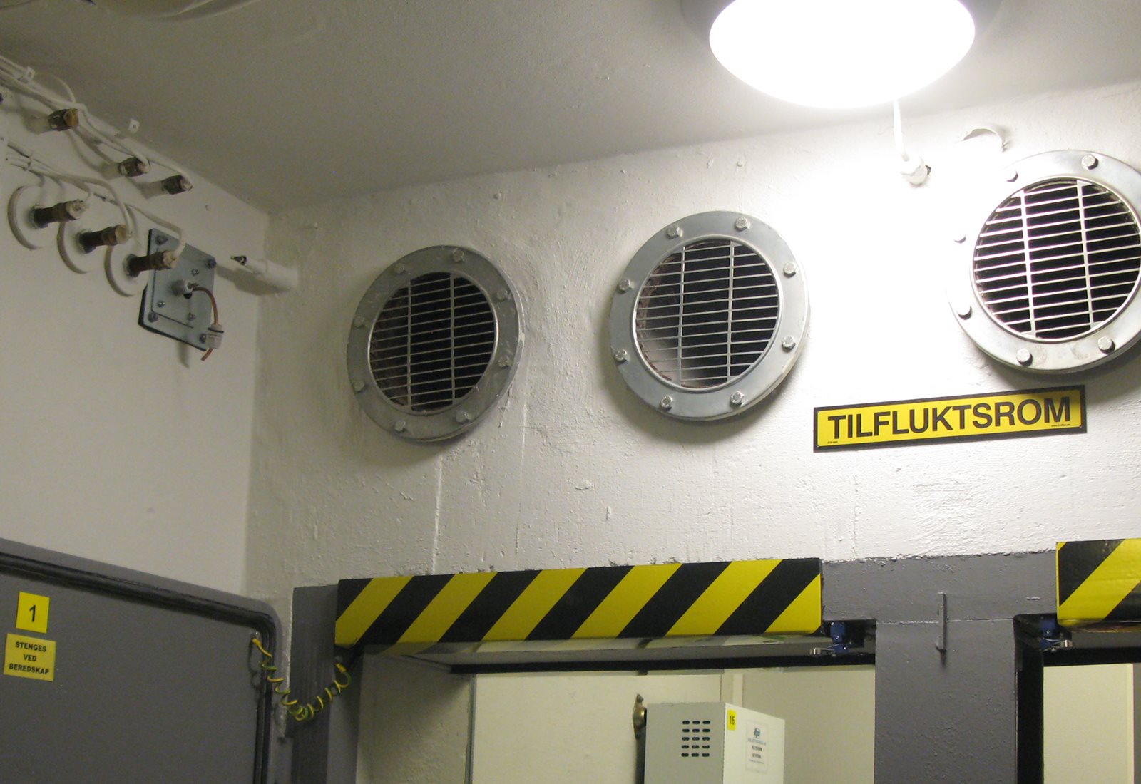 Kjellervegg i mur med lufteventiler og et gult skilt med sort tekst der det står tilfluktsrom.